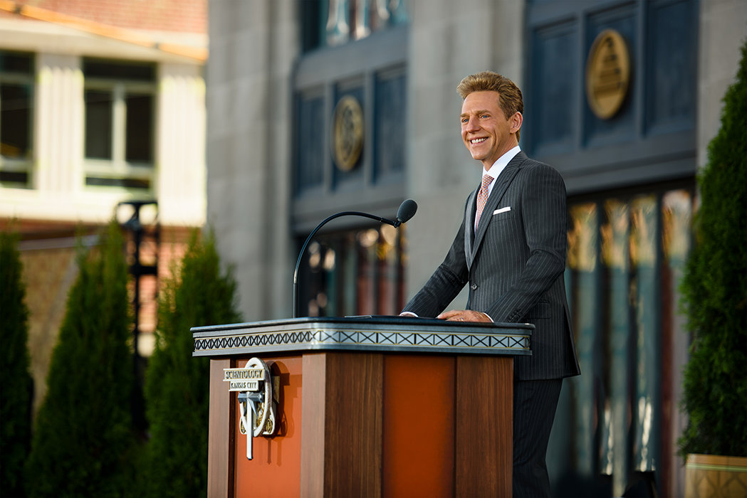 David Miscavige in Kansas City, Missouri, at podium, dark gray suit, smiling